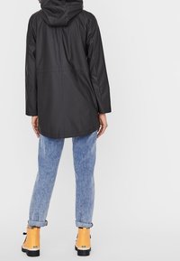 Czarna wodoodporna kurtka z kapturem, o prostym kroju i minimalistycznym designie. Zestawiona z jasnoniebieskimi dżinsami i żółtymi kaloszami.