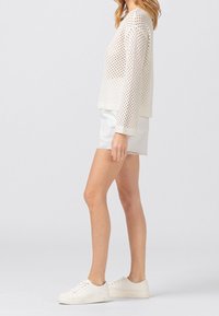 Lekki beżowy sweter z siateczki o luźnym kroju, okrągłym dekolcie, ściągaczach na mankietach i na dole, zestawiony z białą spódnicą dżinsową oraz białymi trampkami.