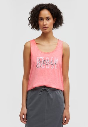 Frau mit lockigem Haar, die ein ärmelloses korallenrosa Top mit Schriftzug und einen grauen Rock mit Kordelzug vor einem schlichten Hintergrund trägt.