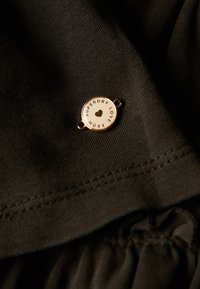 Vêtement en tissu noir avec une étiquette métallique circulaire gravée de "SUPERDRY LOVE", agrémentée d'un symbole de cœur et de bords cousus.