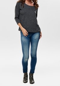Vrouw in een donkergrijze sweater met lange mouwen, gescheurde blauwe skinny jeans en slangenhuid enkellaarsjes voor een effen achtergrond.