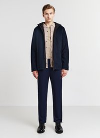 Marineblaue Fleecejacke mit Kapuze, getragen über einem beigen Hemd mit Knöpfen. Schwarze Hose und Schuhe runden das Outfit ab. Glattes, strukturiertes Material.