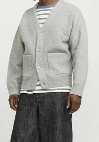 Grå cardigan i ull med V-hals, framfickor och ribbade muddar. Lager på en randig T-shirt. Mörka jeans syns.