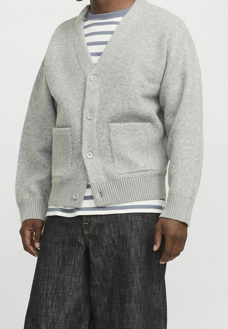 Grå cardigan i ull med V-hals, framfickor och ribbade muddar. Lager på en randig T-shirt. Mörka jeans syns.