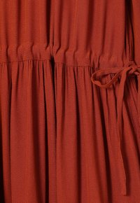 Tissu couleur rouille avec détail froncé à la taille et ceinture en tissu nouée sur le côté droit, mettant en valeur la texture et les plis.