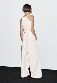 Wijdvallende witte broek met hoge taille en zijzakken, gecombineerd met een aansluitend wit tanktop, getoond vanaf de achterkant.