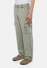 Muž má na sobě vybledlé zelené cargo kalhoty s bočními knoflíkovými kapsami a béžové semišové slip-on boty s přezkami.