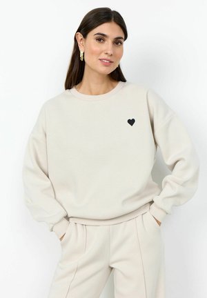 Woman with dark hair wearing beige sweatshirt and matching pants, with small black heart on left chest and gold earrings, standing with hands in pockets.