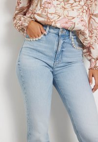 Mujer vistiendo jeans de talle alto azul claro con adornos de perlas en los bolsillos y una blusa floral de manga larga, con la mano en el bolsillo.