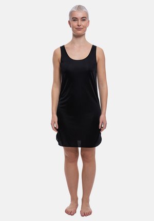 Woman with short blonde hair standing barefoot, wearing a sleeveless black dress with a scoop neckline against a plain white background.