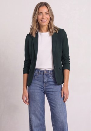 Femme souriante portant un blazer vert foncé, une chemise blanche et un jean bleu, debout devant un fond clair uni.