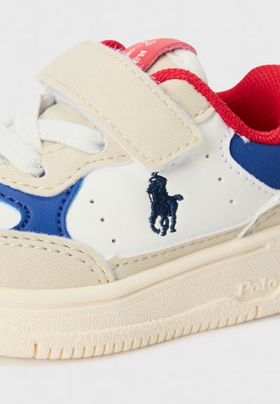 Zapatilla beige y blanca con detalles en azul, que cuenta con una correa de velcro, cordones de textil y un logo bordado en el lateral. Suela de goma.
