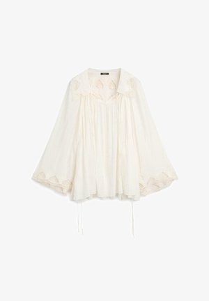Blouse blanche ample avec manches larges, détails en dentelle sur les poignets et le col, et tissu froncé sous l'encolure avec liens coulissants.