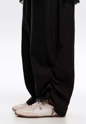 Pantalones negros de pierna ancha con puños fruncidos recogidos, combinados con zapatillas deportivas beige claro con cordones blancos y rojos sobre un fondo blanco.