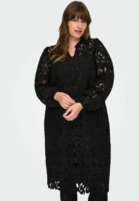 Crna čipkasta haljina s cvjetnim uzorcima i valovitim rubom. Ima duge rukave i V-izrez, izrađena je od teksturiranog materijala.