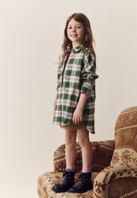 Grünes und weißes kariertes Hemdblusenkleid mit Knöpfen, über einem beigen Rock getragen. Schwarze Schnürstiefel mit gemusterten Schnürsenkeln, pinke Socken.