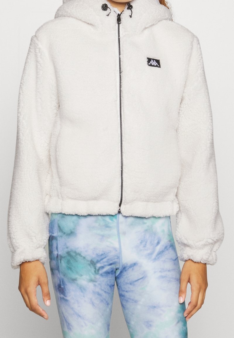 Sudadera blanca y esponjosa con cremallera y capucha, puños elásticos y un parche de logo negro. Combinada con leggins de teñido anudado en azul y verde.