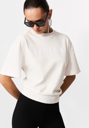 Fehér, rövidített póló "LAMANIA" logóval, texturált betűkkel. Rövid ujjú, kerek nyakkivágással. Fekete leggingsszel párosítva.