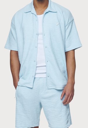 Man wearing light blue textured short-sleeve button-up shirt over white t-shirt and matching textured shorts.