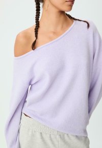 Hellblau-lila, übergroßer Pullover mit einem einseitigen Off-Shoulder-Design und langen Ärmeln, aus weichem Stoff gefertigt. Getragen mit grauen, elastischen Bundhosen.