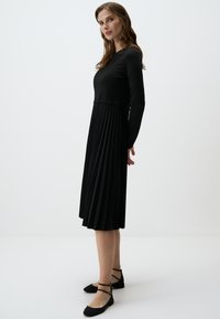 Robe noire à manches longues, corsage ajusté et jupe plissée. Associée à des ballerines noires avec des brides autour de la cheville et des accents dorés.