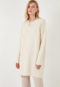 Pull crème à manches longues à côtes avec une coupe décontractée et des fentes latérales. Il présente un col rond et un tissu texturé, associé à un pantalon neutre.