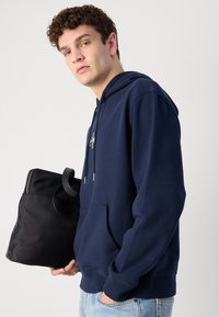 Felpa blu marine con tasca frontale, cappuccio con cordino e dettaglio del logo. Il modello tiene una borsa tote in tessuto nera con zip e manico.