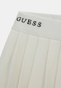 Jupe plissée crème avec une ceinture côtelée ornée du mot « GUESS » brodé en noir. Texture douce et design ajusté.