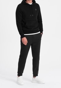 Mann in schwarzem Kapuzenpullover und passenden schwarzen Jogginghosen mit weißen Sneakers, steht mit den Händen in den Taschen vor einem weißen Hintergrund.