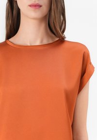 Mujer con una blusa naranja de manga corta y cuello redondo, sobre un fondo blanco liso.