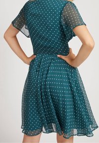 Nainen, joka on pukeutunut teal-väriseen lyhythihaiseen mekkoon, jossa on valkoisia pilkkuja, kädet lanteilla, kuvattuna selkäpuolelta yksinkertaista taustaa vasten.