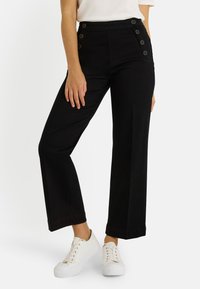 Pantalones negros de tiro alto con diseño de pierna ancha y cinco botones decorativos en el lado izquierdo. Combinados con zapatillas blancas.