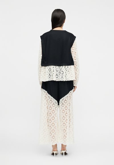 Γυναίκα με μαύρο μπλουζάκι χωρίς μανίκια πάνω από λευκό μακρυμάνικο δαντελένιο πουκάμισο και ταιριαστά δαντελένια παντελόνια με φαρδιά μπάτσα, στέκεται σε απλό φόντο, από την πίσω όψη.