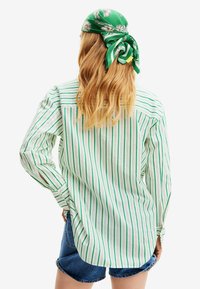 Persona con lunghi capelli biondi che indossa una maglietta a righe verdi e bianche, pantaloni corti di denim blu e un foulard floreale verde legato dietro la testa.