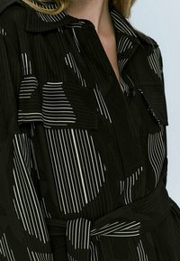 Giacca nera con strisce bianche e pattern astratti. Presenta un design con bottoni, colletto e cintura in vita. Due tasche sul petto.