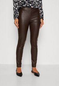 Mujer vistiendo pantalones ajustados de cuero marrón oscuro, tacones slingback negros de punta afilada y una blusa con un estampado blanco y negro.