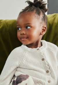 Striktrøje i lys beige med en flæsekant og træknapper, parret med mønstrede bukser. Barn med struktureret hår og et subtilt smil.