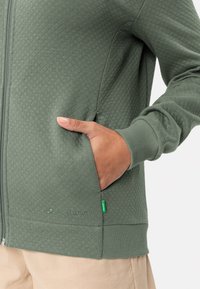 Giacca verde con zip in un tessuto trapuntato, con tasche laterali e polsini a coste. Include un piccolo etichetta verde vicino all'orlo.