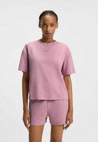 Donna che indossa un completino coordinato composto da una t-shirt oversize mauve e dei pantaloni shorts, con la scritta "HUGO" sul petto, in piedi su uno sfondo bianco uniforme.