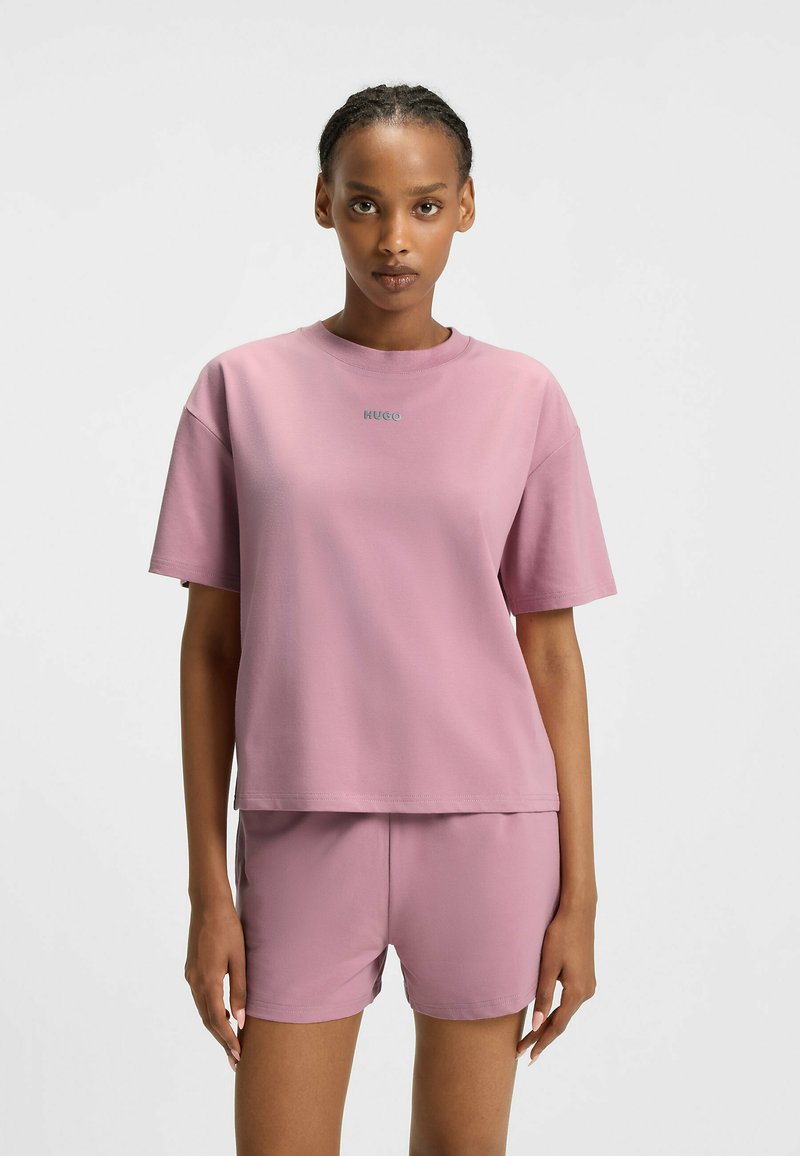 Donna che indossa un completino coordinato composto da una t-shirt oversize mauve e dei pantaloni shorts, con la scritta "HUGO" sul petto, in piedi su uno sfondo bianco uniforme.