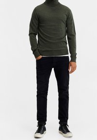 Maglione verde a collo alto, design aderente, polsini e orlo a costine, abbinato a jeans neri e sneaker alte nere.