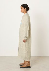 Dolga, svetlo siva, rebrasta kardigan s prenapoljenimi rokavi, zapiranjem na gumbe in stranskimi razpoki; v kombinaciji z bež gležnjarji z okrasnimi elementi.