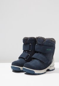 Botas infantiles azul marino con forro de piel gris, dos correas ajustables y suelas de goma texturizadas, con sutiles detalles en verde y logo de la marca.
