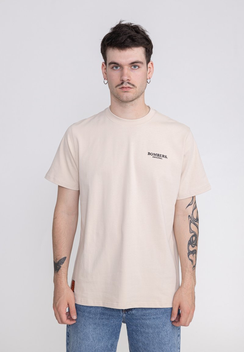 Beige Baumwoll-T-Shirt mit kurzen Ärmeln und rundem Halsausschnitt, das ein kleines schwarzes "BOMBERS"-Logo auf der linken Brustseite trägt.