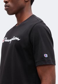 Άνδρας φοράει μαύρο T-shirt Champion με λευκό και κόκκινο λογότυπο στο στήθος και μικρό λογότυπο στο αριστερό μανίκι σε μονόχρωμο φόντο.