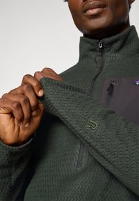 Pull en polaire texturée verte avec col montant, fermeture éclair et poche poitrine noire. Présente un écusson logo sur la manche.