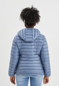 Lichtblauwe gewatteerde jas met een capuchon, voorzien van horizontaal gestikte panelen. Soepele, glanzende stof en elastische manchetten. Draagt over lichte spijkerbroeken.