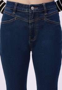 Donkerblauwe denim jeans met een hoge taille, voorkwabben en contrasterende oranje stiksels langs de naden. Zachte textuur, knoopsluiting.