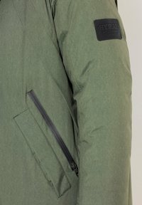 Chaqueta de tela verde con una textura suave, que cuenta con un bolsillo negro con cremallera y un parche con el logo en la parte superior de la manga. Diseño ajustado.