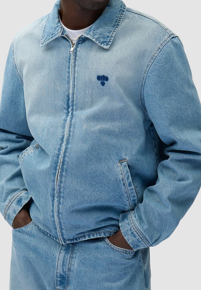 Uomo con giacca di denim azzurro chiaro e jeans abbinati, mani nelle tasche, la giacca presenta un piccolo logo scuro "arfe" sul petto.